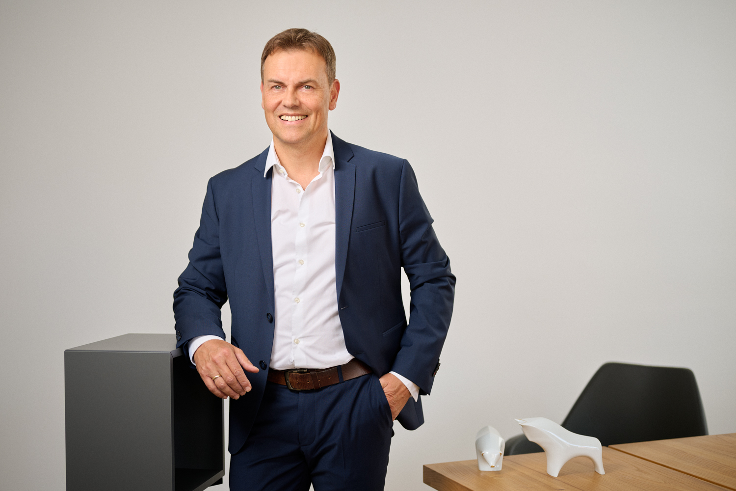 Portrait von Wolfgang Altmann: Ein Mann mittleren Alters mit braunem, kurzen Haar, in dunkleblauen Anzug und weißem Hemd steht lachend in einem modernen Büro. Eine Hand ruht auf einem dunklen Möbelstück, im Hintergrund ein Tisch mit weißen Stier- und Bärfiguren.